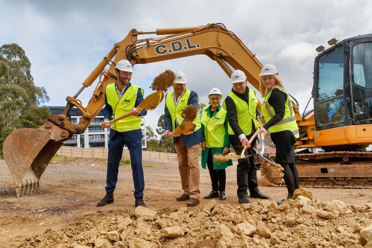 Photograph of people on construction site, turning soil with digger in the background